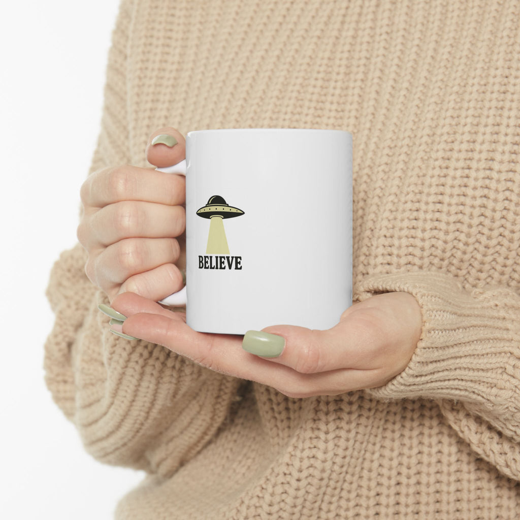 Person holding a white mug with a UFO design and 'BELIEVE' text, wearing a beige sweater.